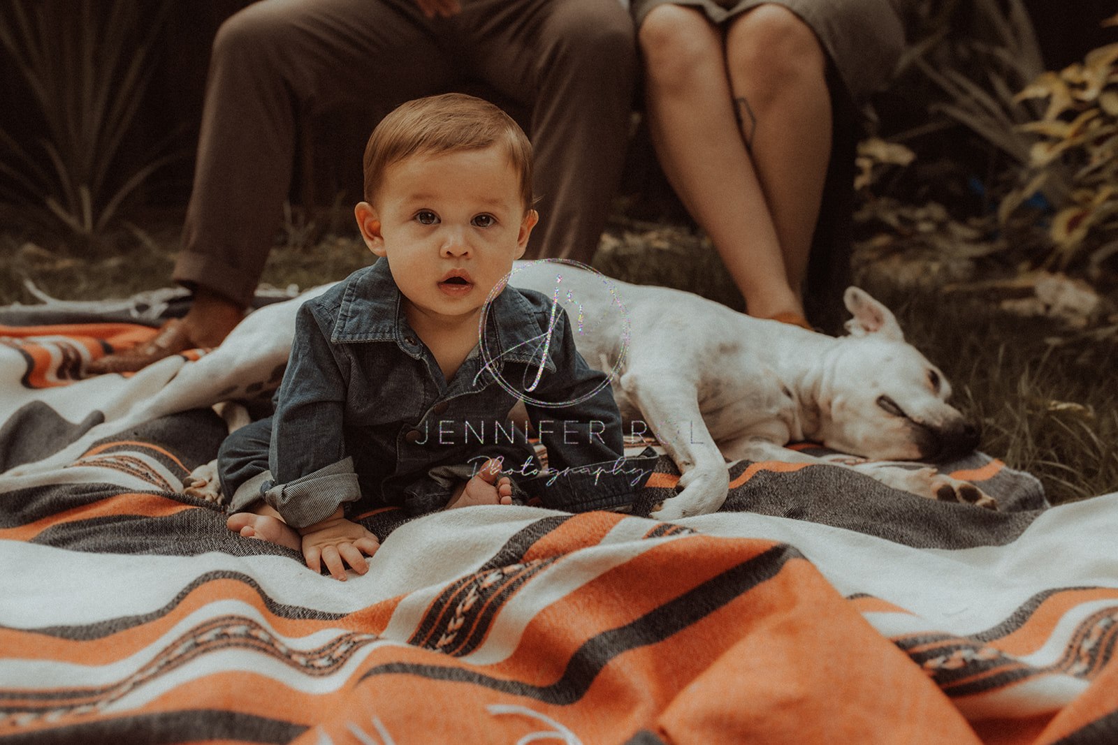 A toddler boy in denim onesie plays on an orange picnic blanket in front of mom, dad and their sleeping dog after visiting baby stores in Missoula