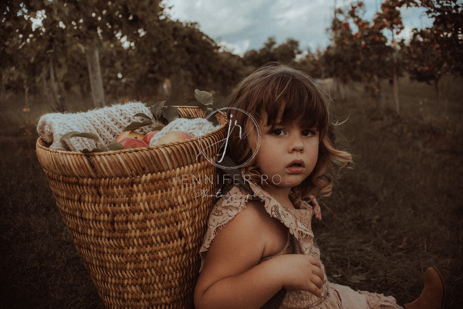 A toddler girl sits on in an apple orchard wearing a wicker basket full of apples and a blanket