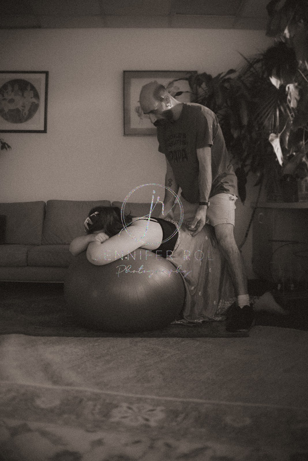 A woman kneels over an exercise ball with her husband helping while preparing for birth in a living room as guided by Missoula Midwives