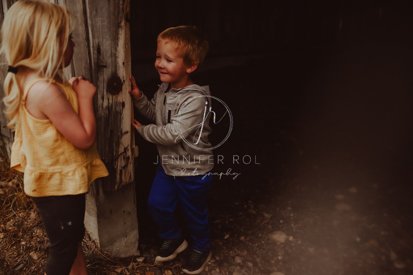 A young boy plays with his toddler sister around a rustic wood wall