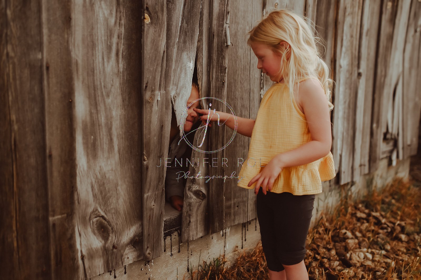 A toddler girl in a yellow shirt boops her brother through a rustic wall after visiting pediatricians in Missoula