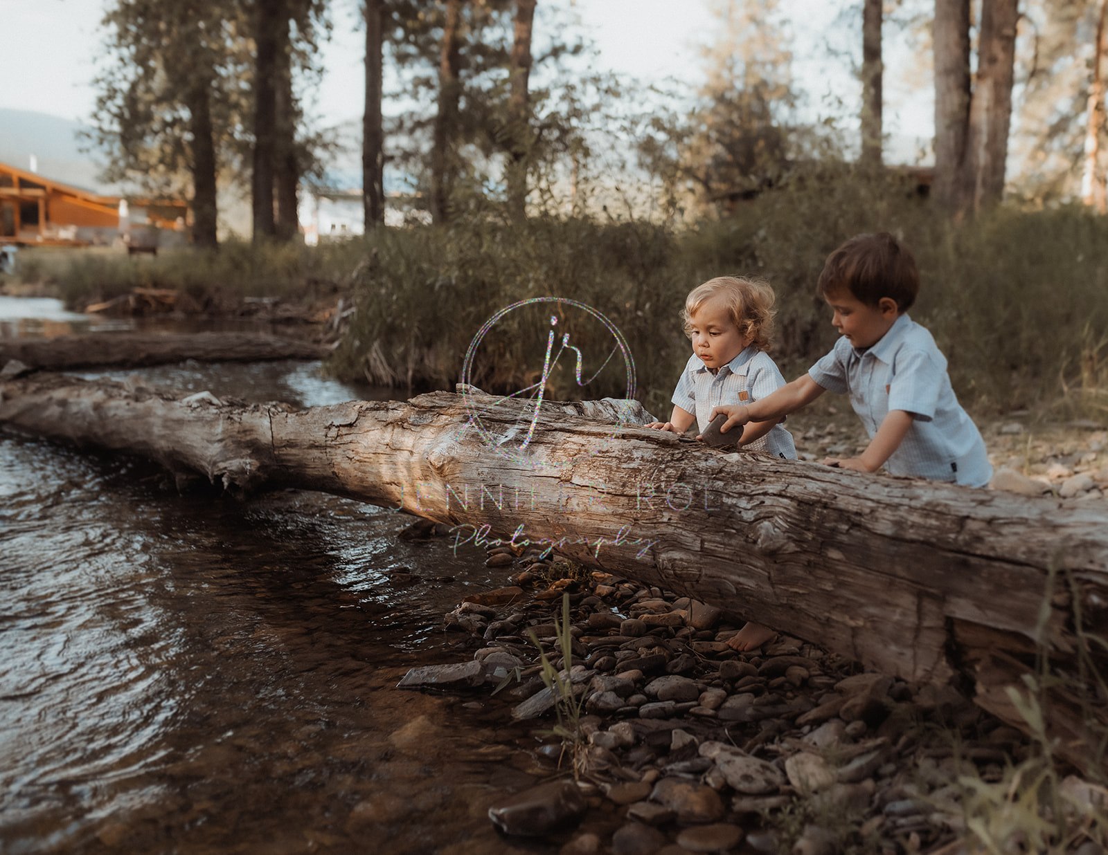 Toddler brothers in matching blue shirts play with rocks on a creekside log before visiting Carousel for Missoula