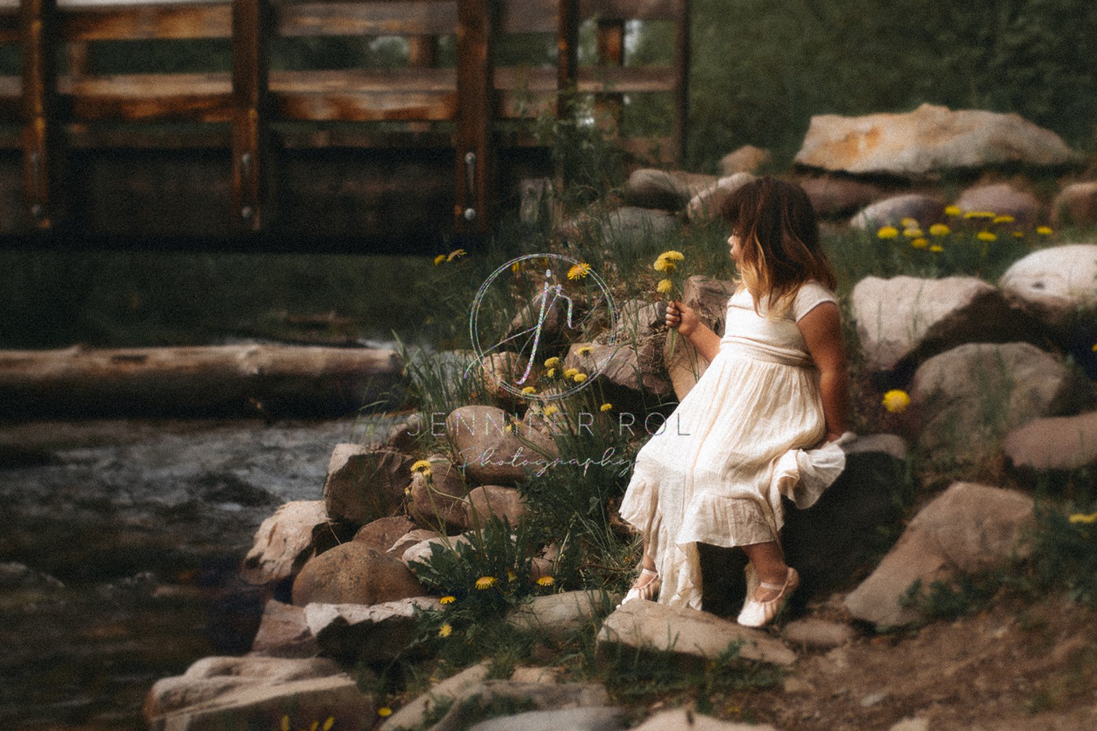 A young toddler girl in a white dress sits on some creekside rocks exploring yellow wildflowers