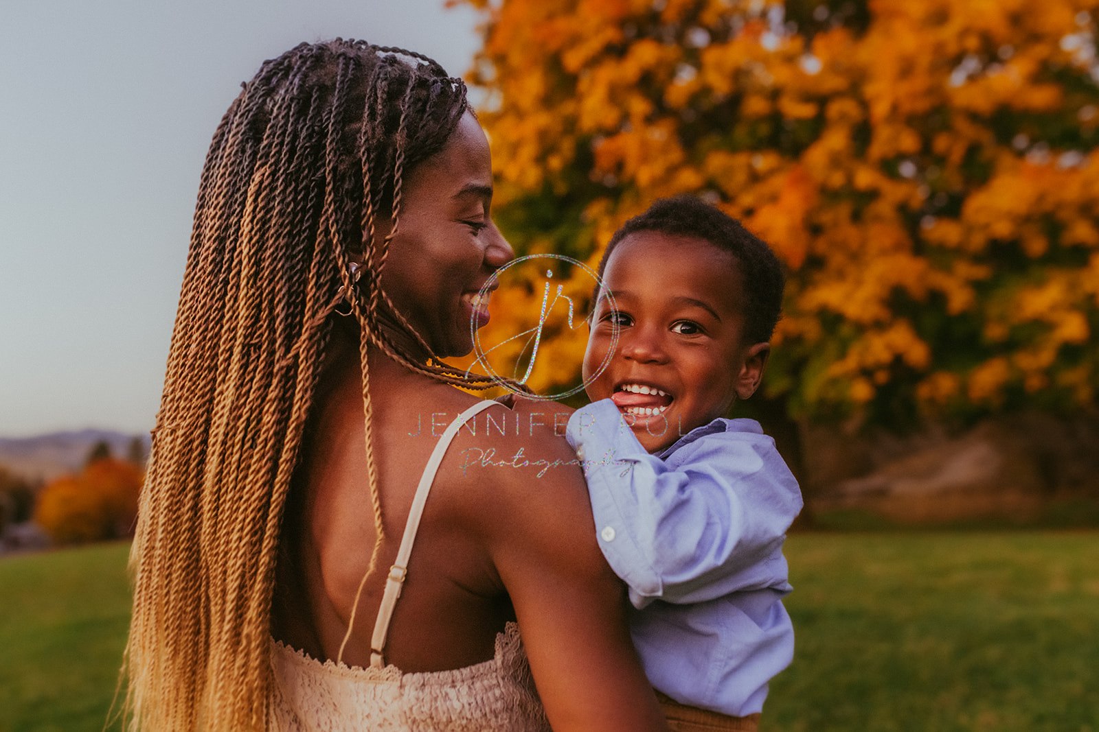 A toddler boy giggles with his tongue out while sitting in mom's arms in a park at sunset