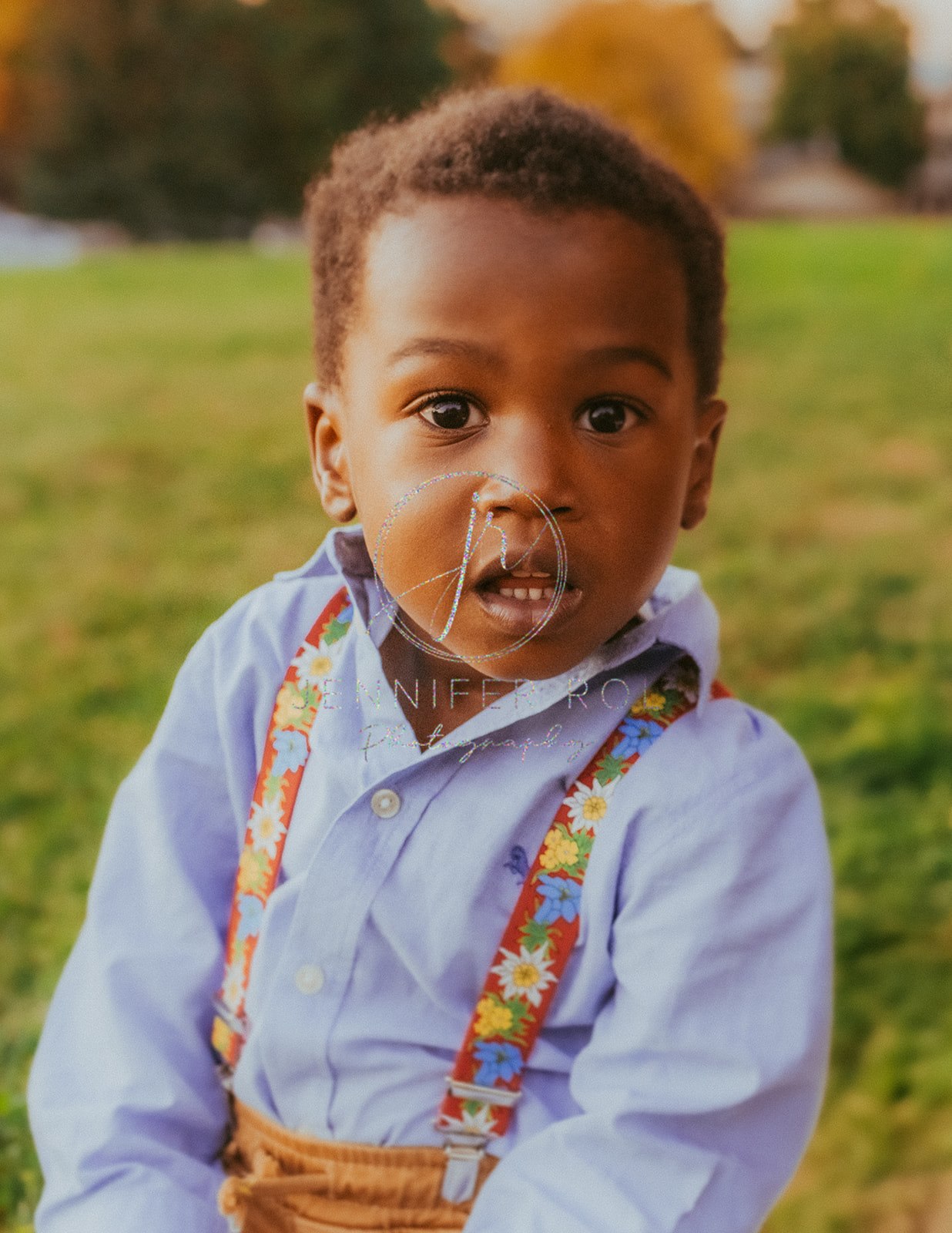 A toddler boy in a blue shirt and flower suspenders plays in a lawn at sunset before visiting Missoula playgrounds