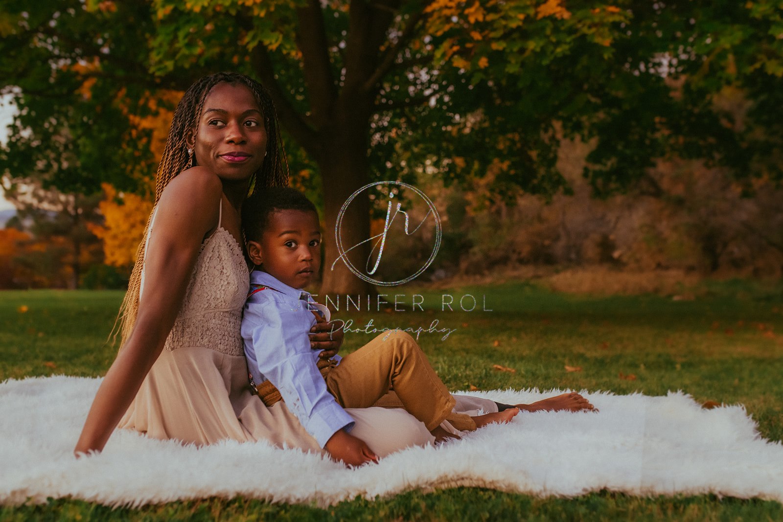 A mom in a beige dress sits on a fur blanket in a park with her toddler son in her lap before visiting Missoula playgrounds