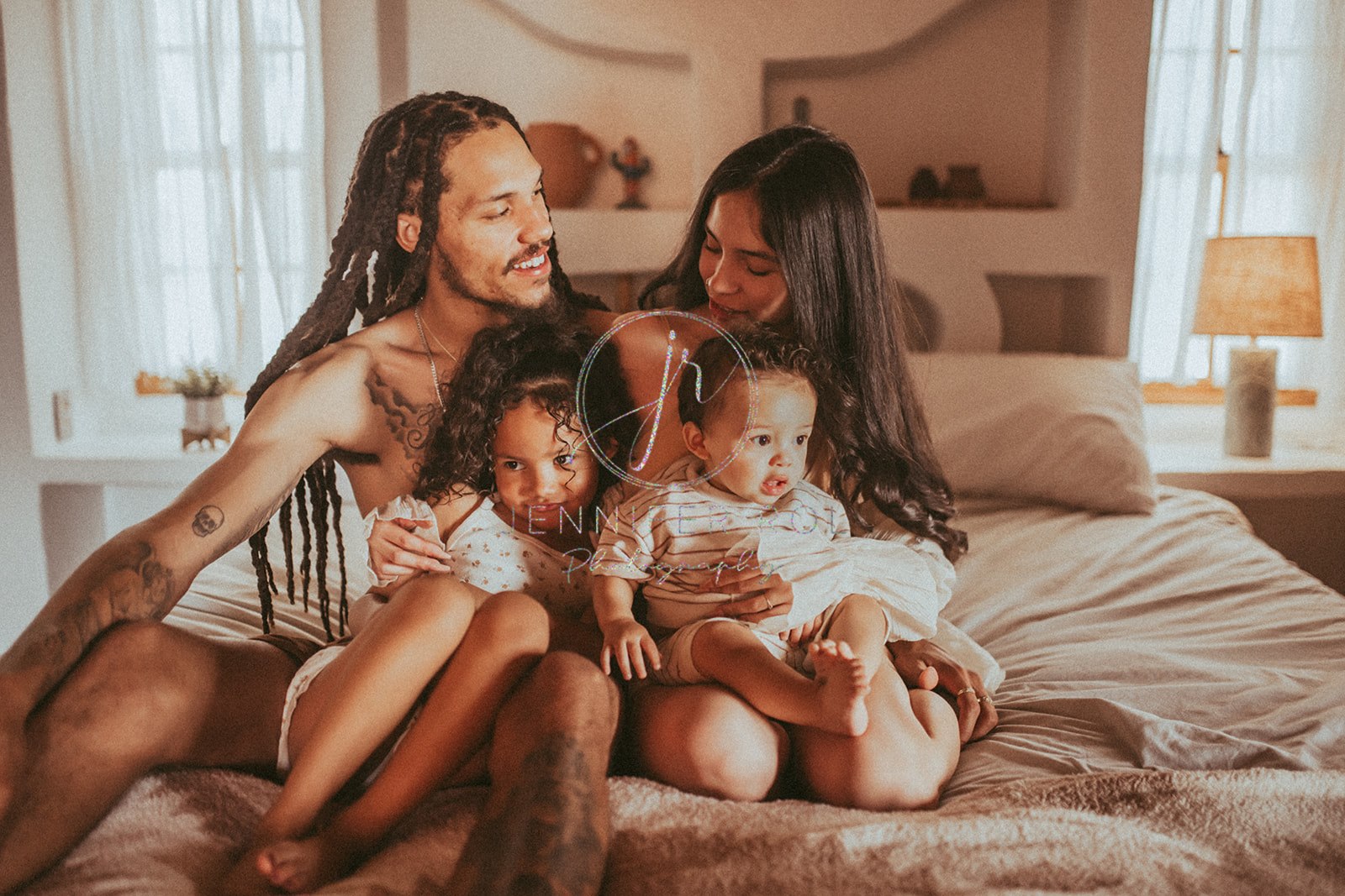 A happy mom and dad sit on a bed with their 2 young children in their laps