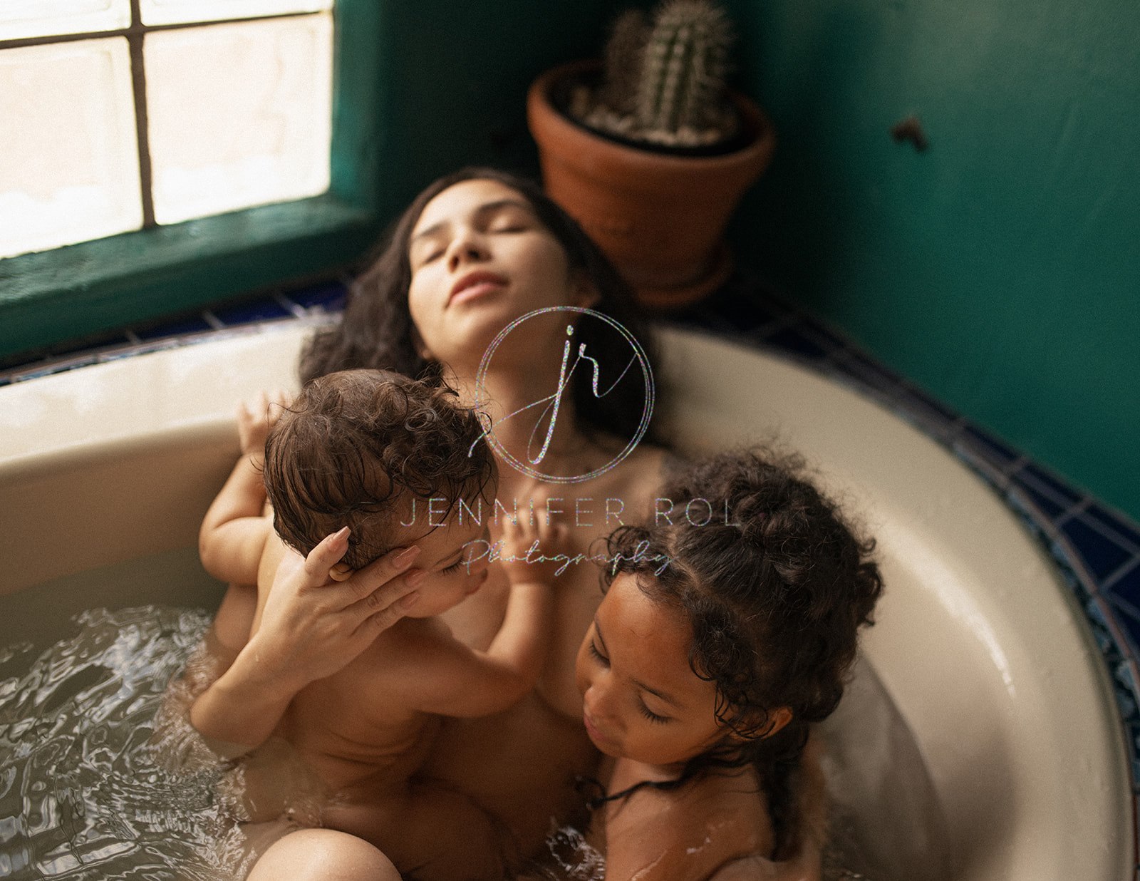 A mom cuddles with her infant and toddler daughter in a bathtub under a window