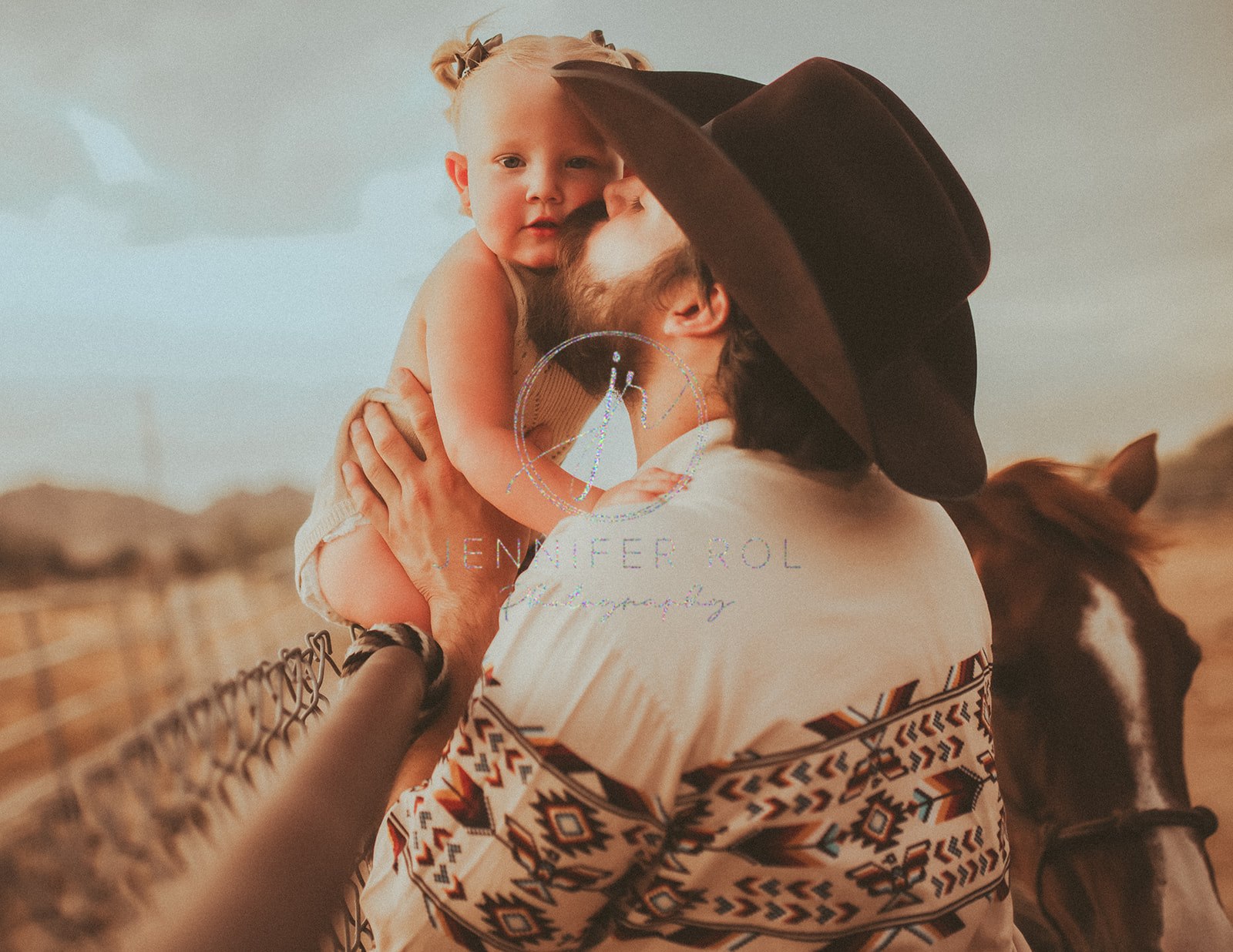 A toddler girl sits on a fence as dad kisses her cheek in a cowboy hat at sunset with a horse