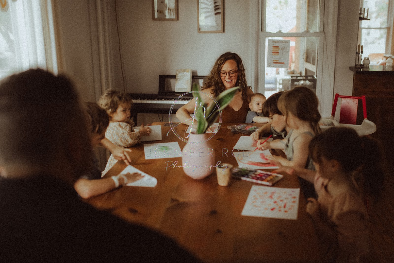 A laughing mom sits with a baby in her lap at a table as her 5 other children color and paint before visiting family restaurants in Missoula, MT