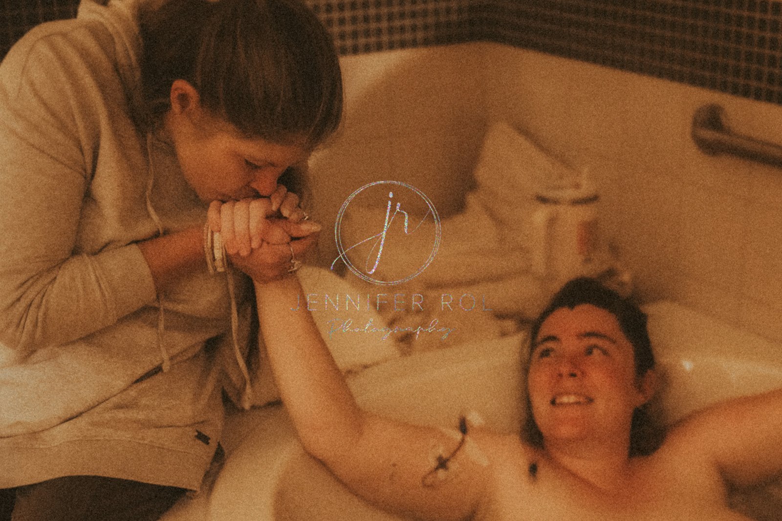 A mom in labor lays in a tub as her partner kisses her hand while guided by Sacred Roots Doula