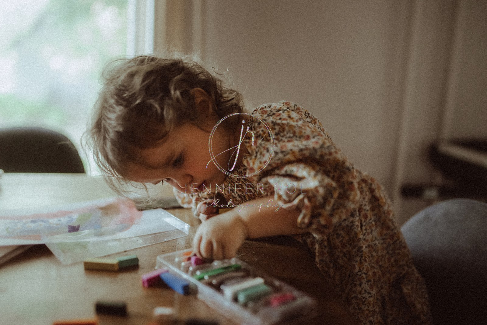 A young toddler in a dress colors with paint supplies at a table and picks a new color from the tray