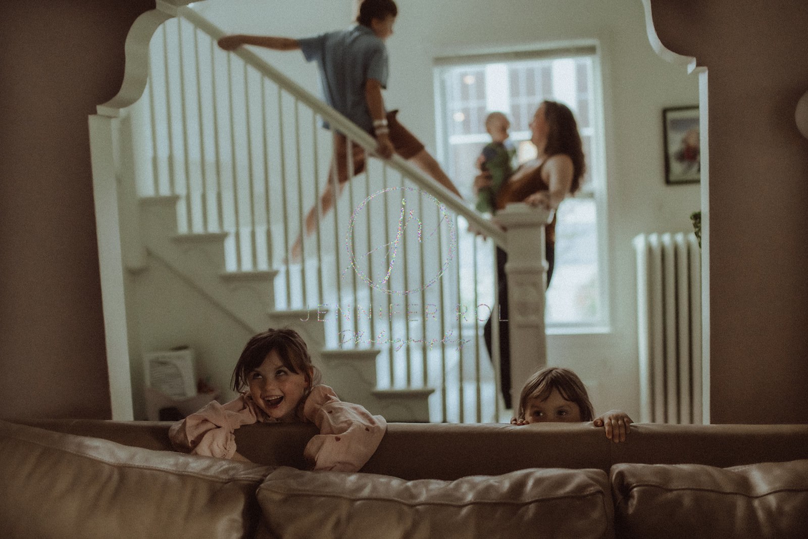 Two sisters play behind a couch as their older brother climbs on the stairs