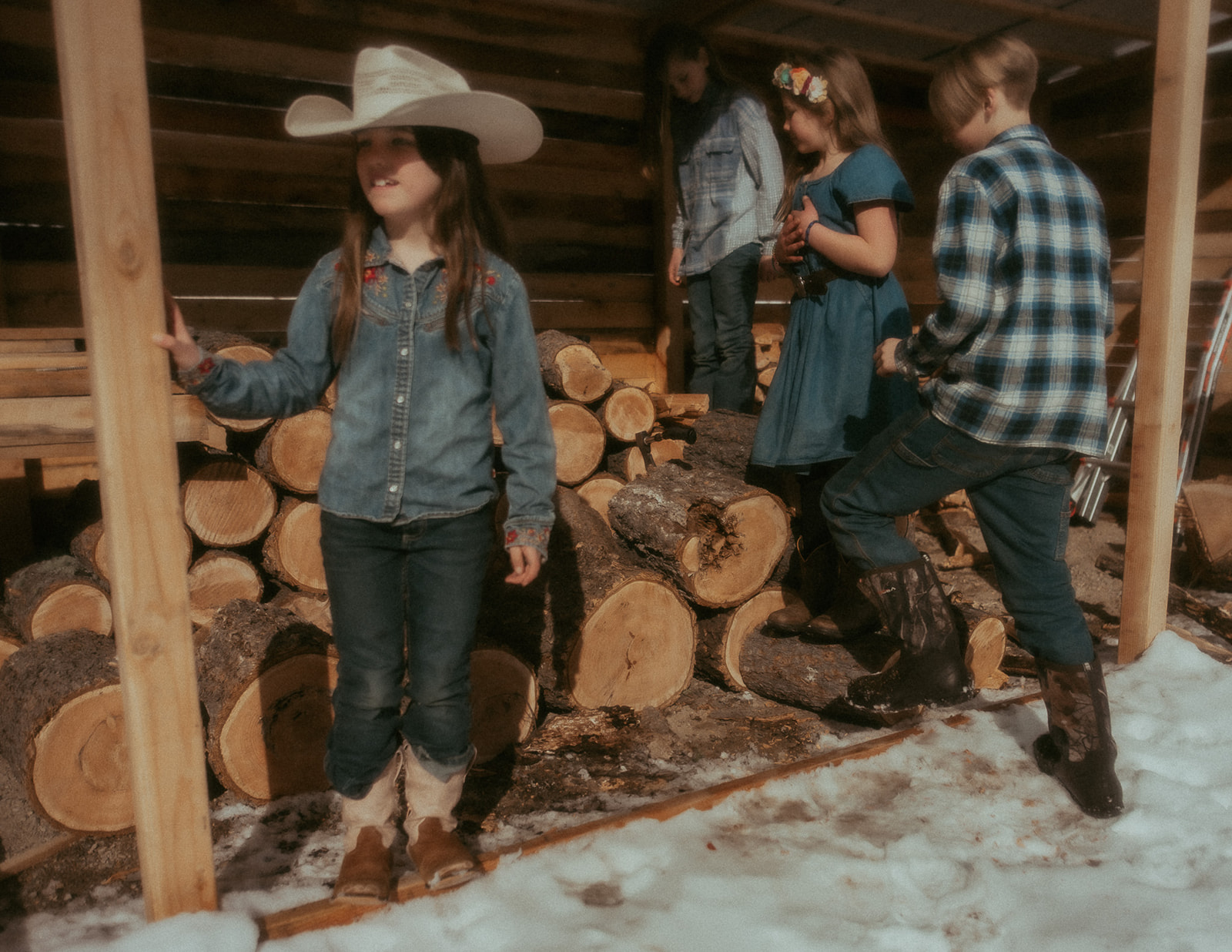 Four siblings play on a pile of wood under a covering on a snowy day in denim and boots after visiting BASE Missoula