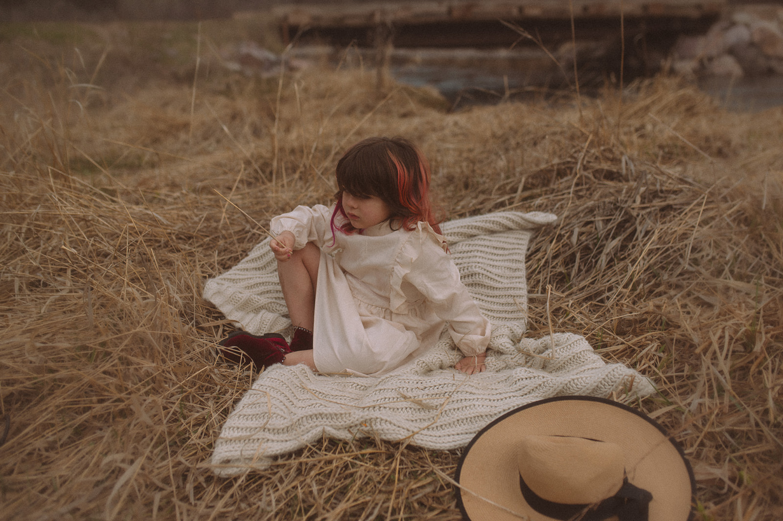 A toddler girl in a white dress sits on a picnic blanket on a pile of hay