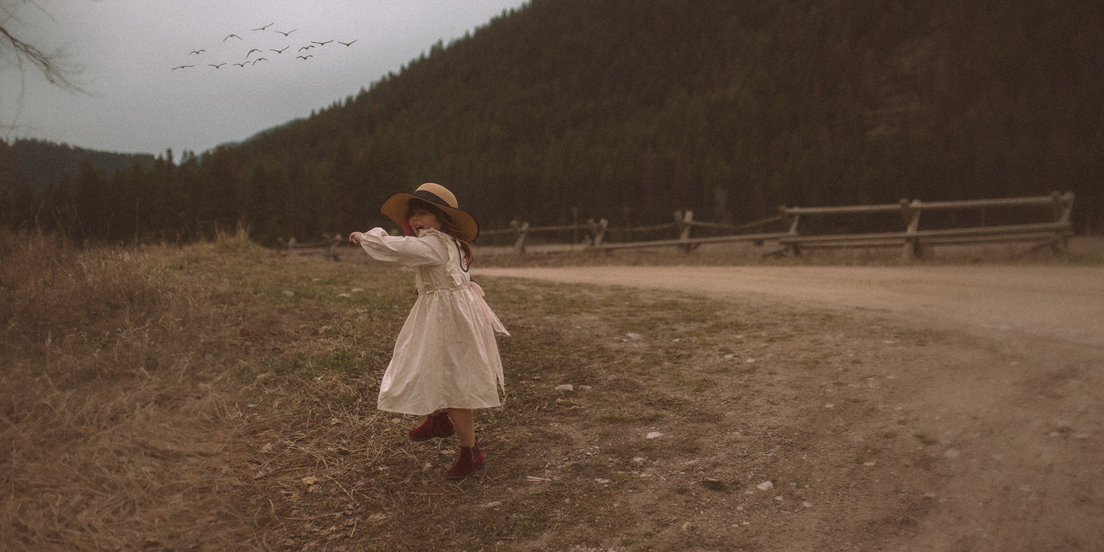 A young girl in a large hat and white dress runs in a dirt road as birds fly over after visiting Dragon Hollow Missoula