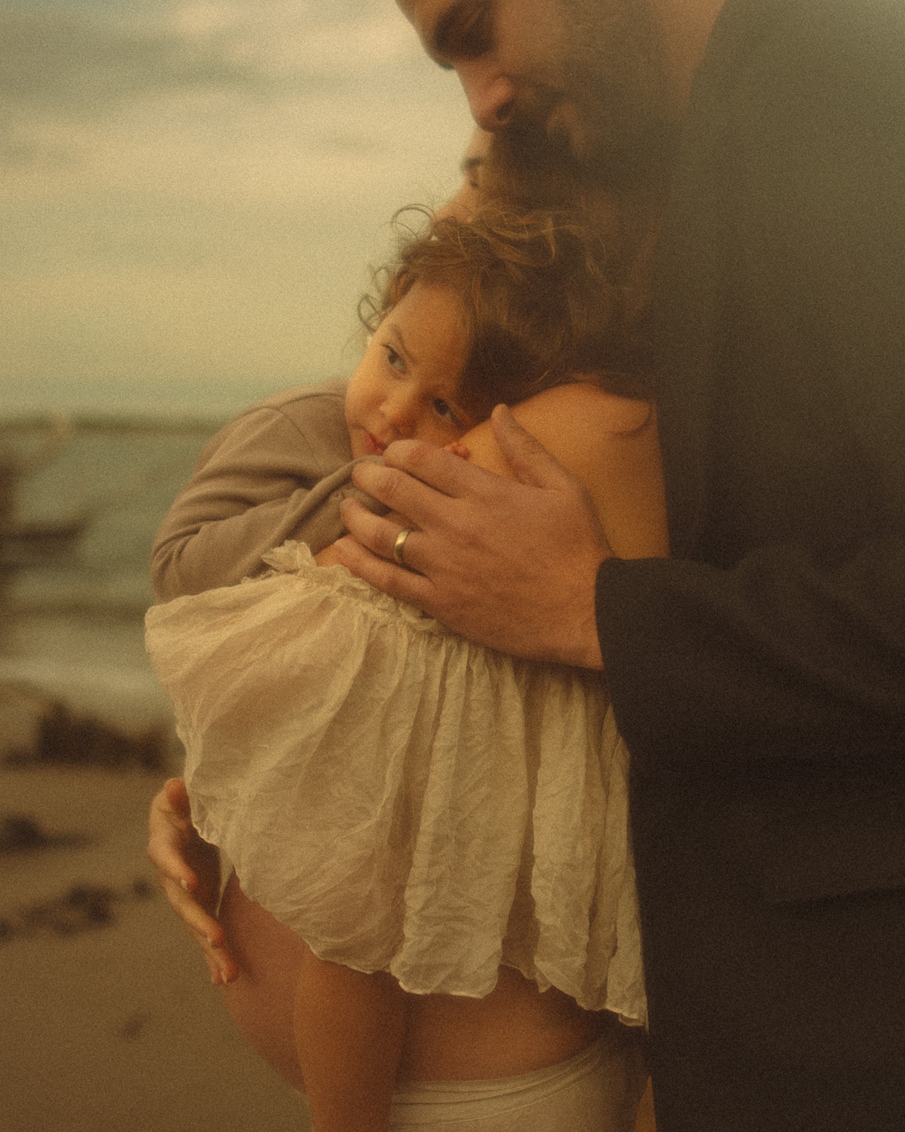 An infant snuggles on mom's shoulder on a beach with dad hugging them after visiting Bright Minds Kids Zone