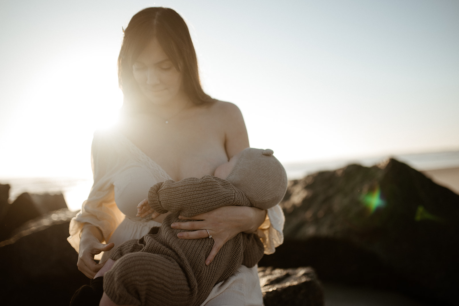 A new mom breastfeeds her newborn on a beach at sunset while sitting on large rocks in a white dress after visiting Butterscotch Babies