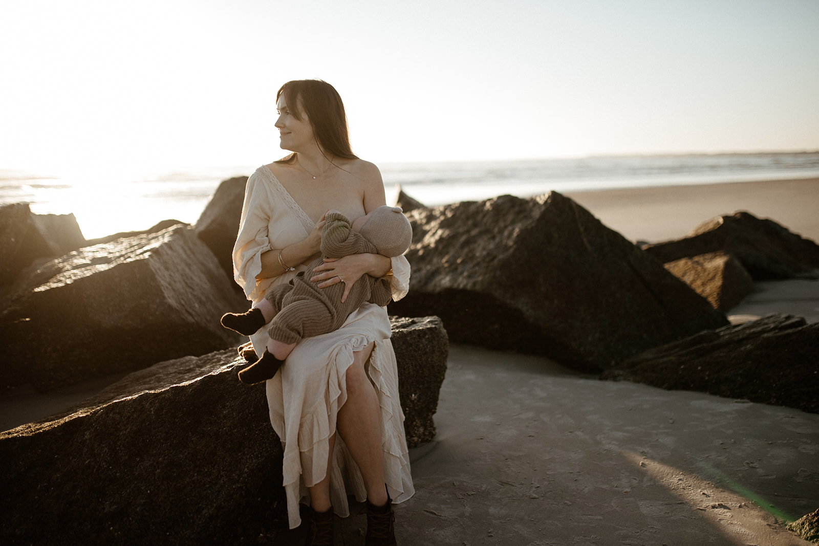 A happy mother sits on a rock on the beach breastfeeding her infant in a white dress at sunset after visiting Butterscotch Babies
