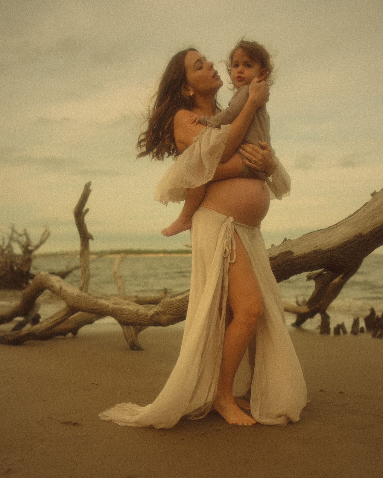 A pregnant mother leans in to kiss her toddler sitting on her bump on a beach at sunset