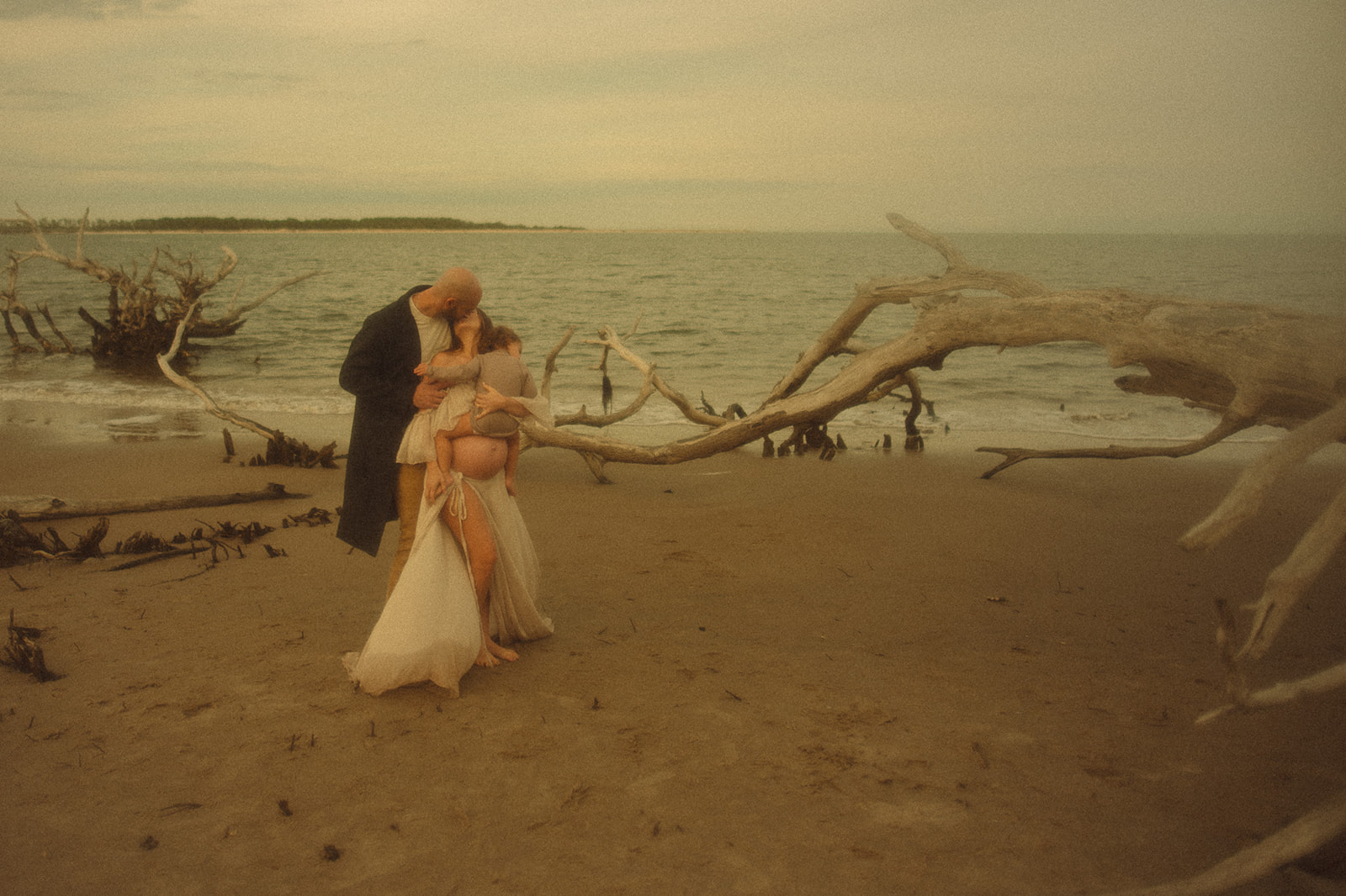 Expecting parents kiss on a beach while their infant sits on mom's bump after visiting Clark Fork Yacht Club