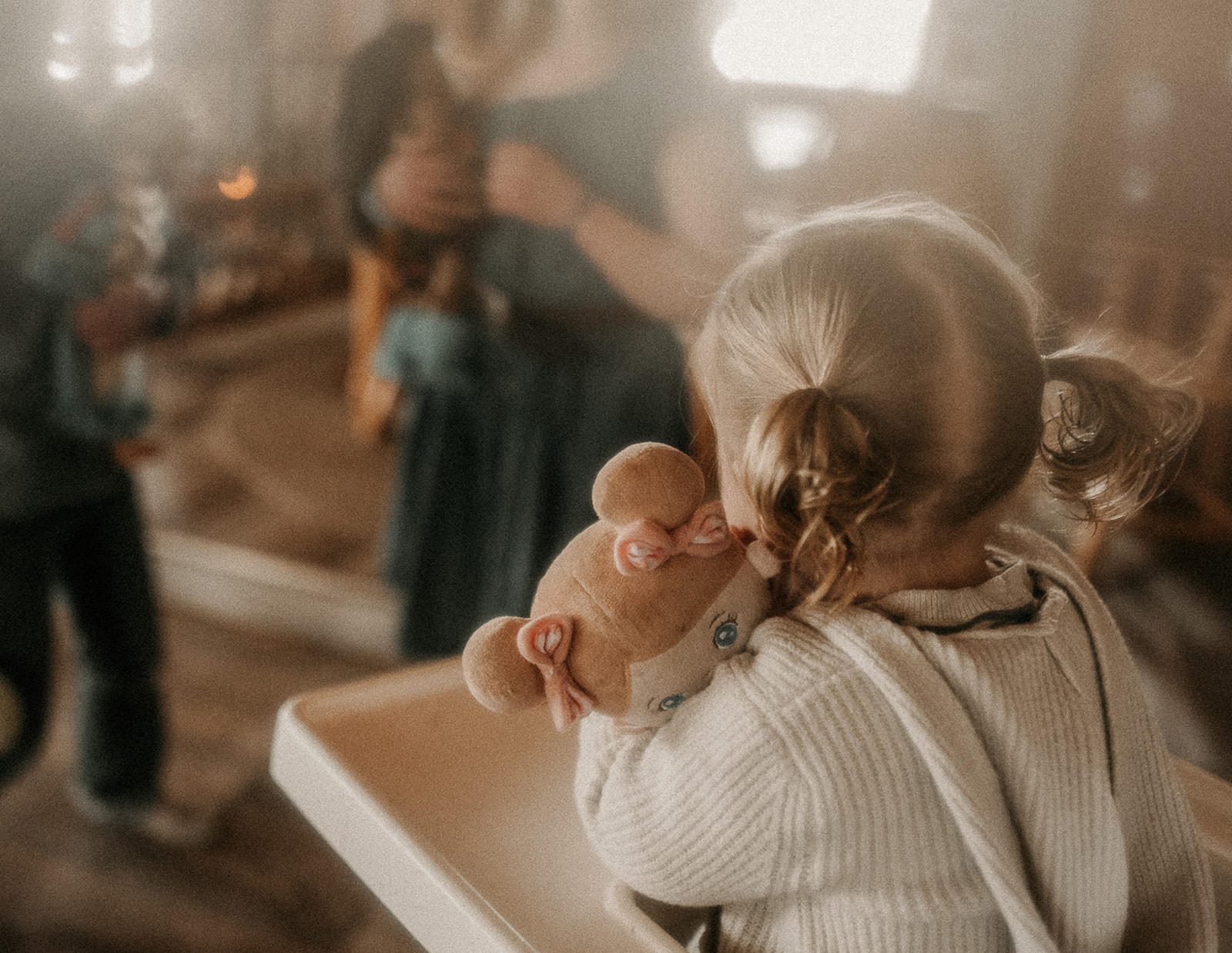 A toddler girl in a white sweater watches mom and dad while clutching her stuffed doll in her high chair after visiting Garden City Ballet