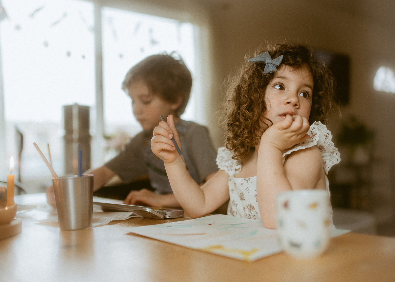 A toddler girl looks over her shoulder while painting at the kitchen table in a white dress with brother behind her
