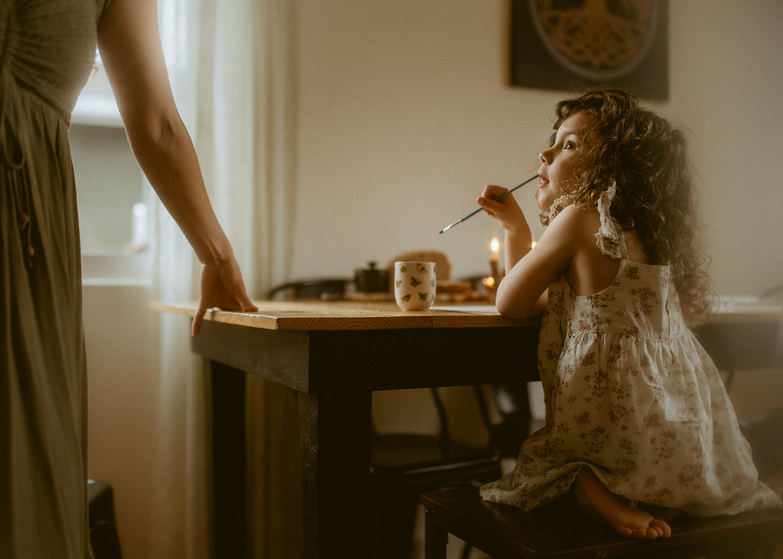 A toddler girl in a floral print dress shows mom her painting at the kitchen table
