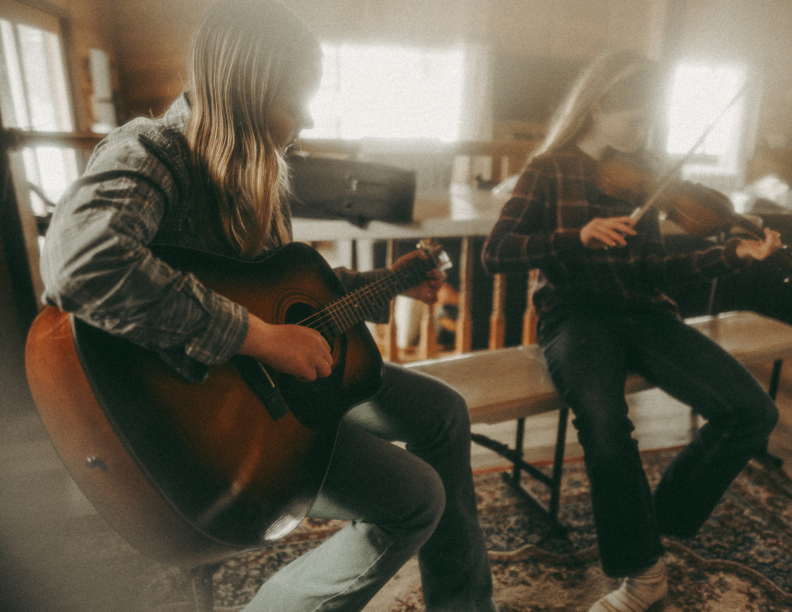 Two sisters in jeans and plaid shirts play violin and guitar on a bench in their home before visiting Farmers Markets in Missoula