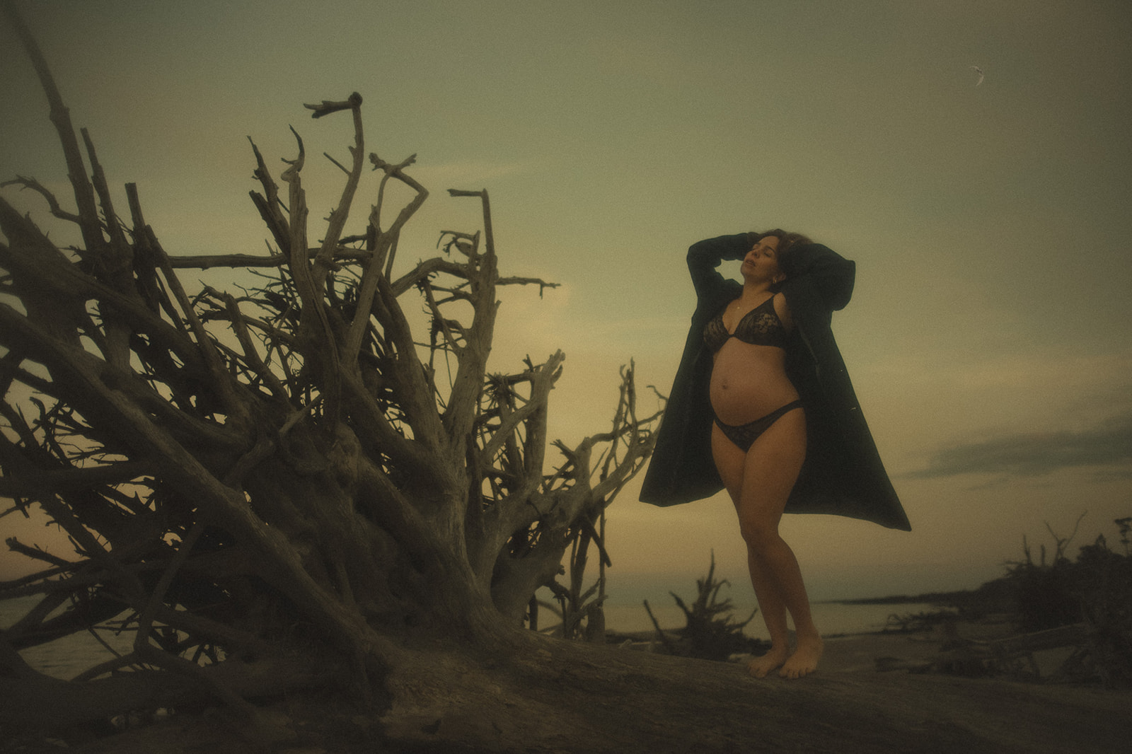 A mom to be in her underwear and a coat basks in the sunset by a scraggly driftwood by the water after visiting Missoula OBGYN Specialists