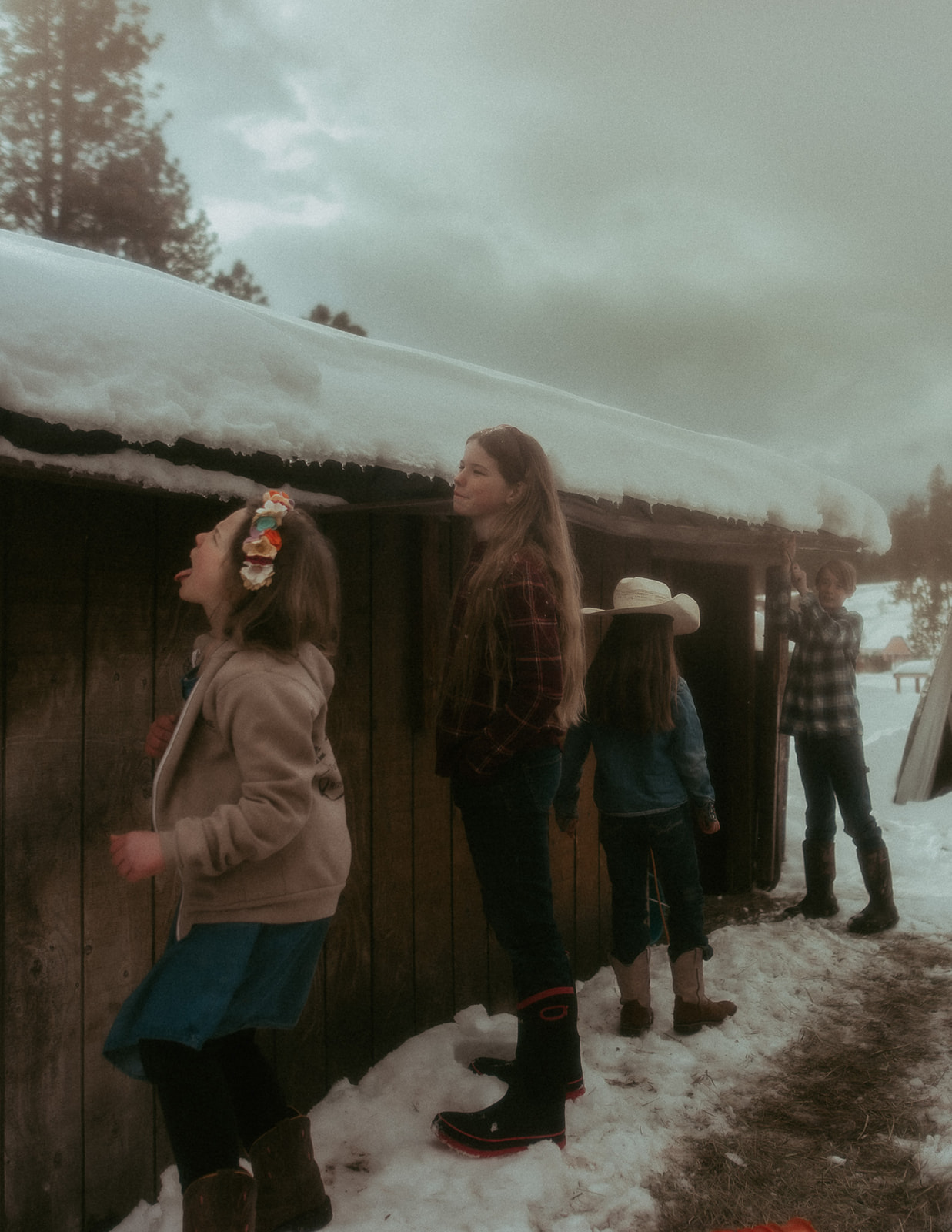 Four siblings play with the snow melting of the roof of a shed in boots and jeans before visiting Missoula PaddleHeads