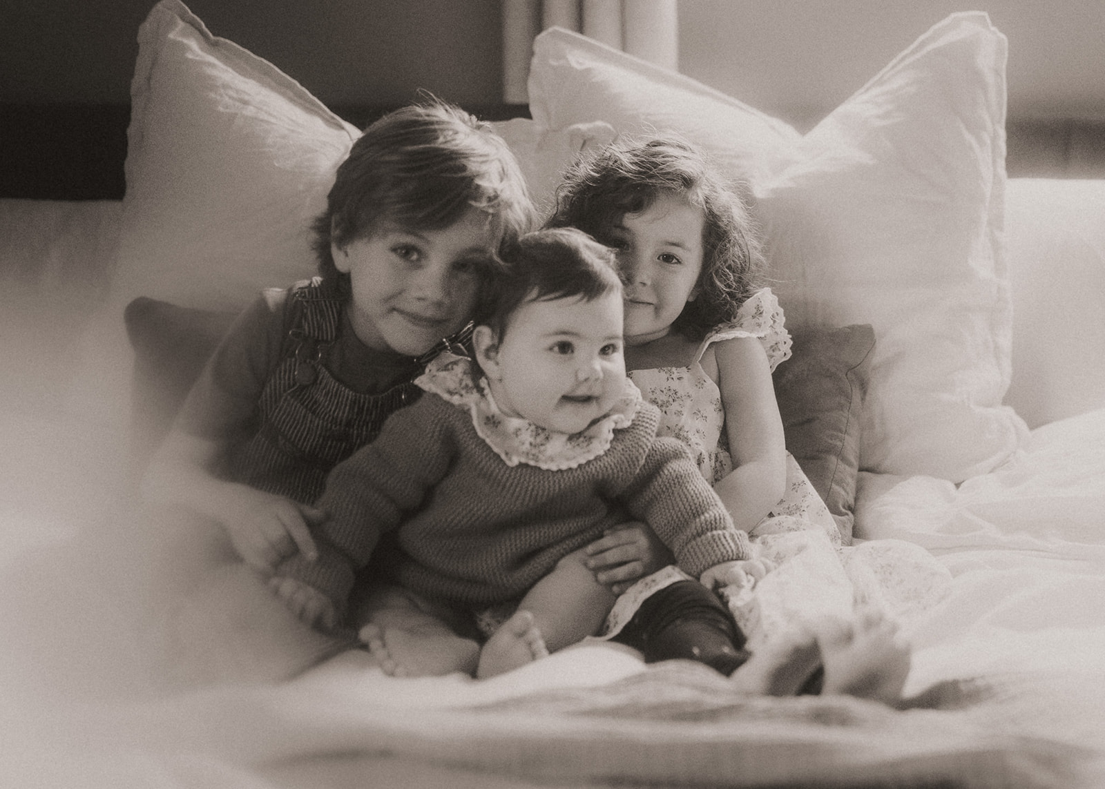A black and white image of three young siblings hugging on a bed