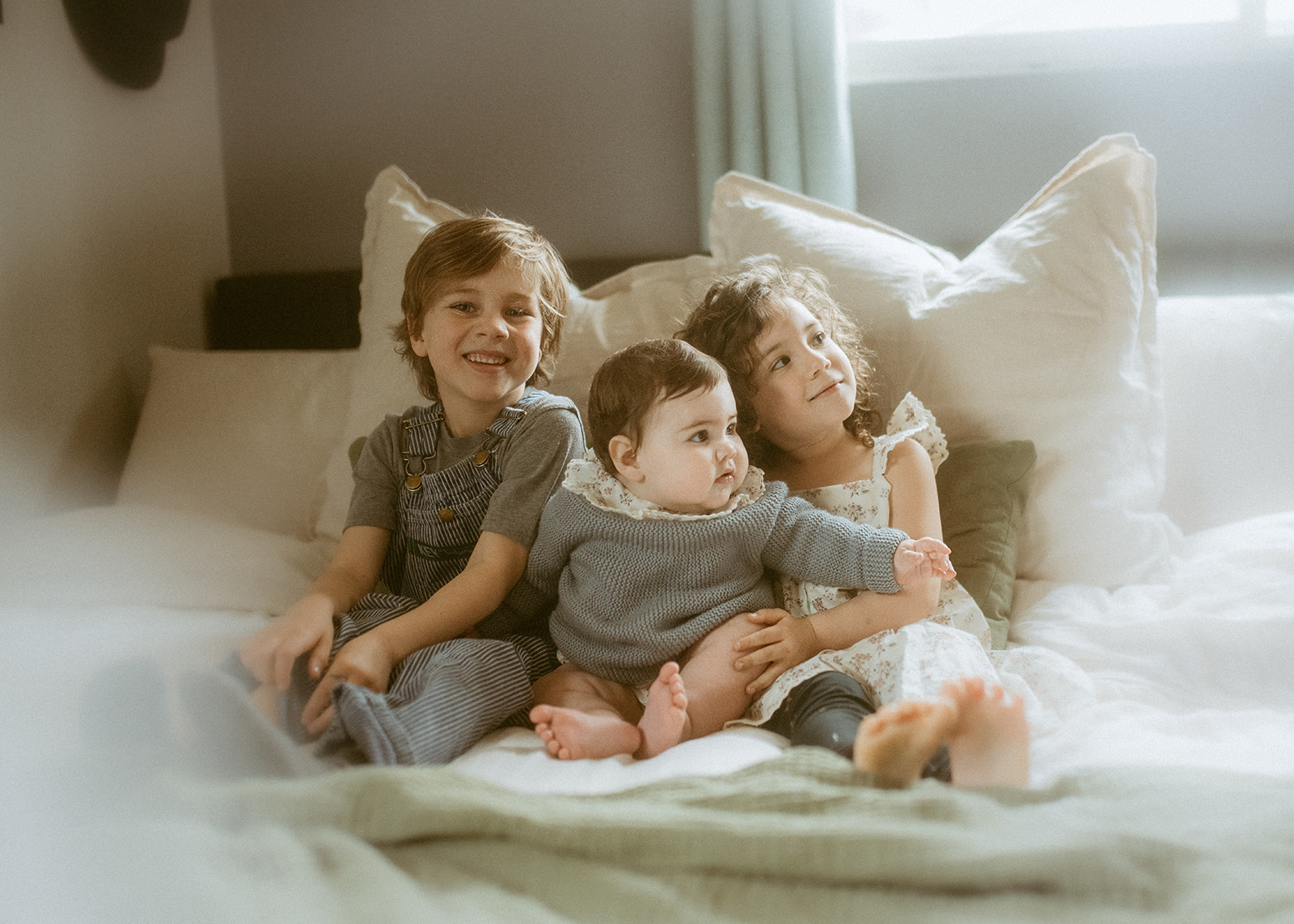 Three happy siblings sit on a bed smiling in overalls, a knit sweater and a floral dress before visiting Roots Acro Sports