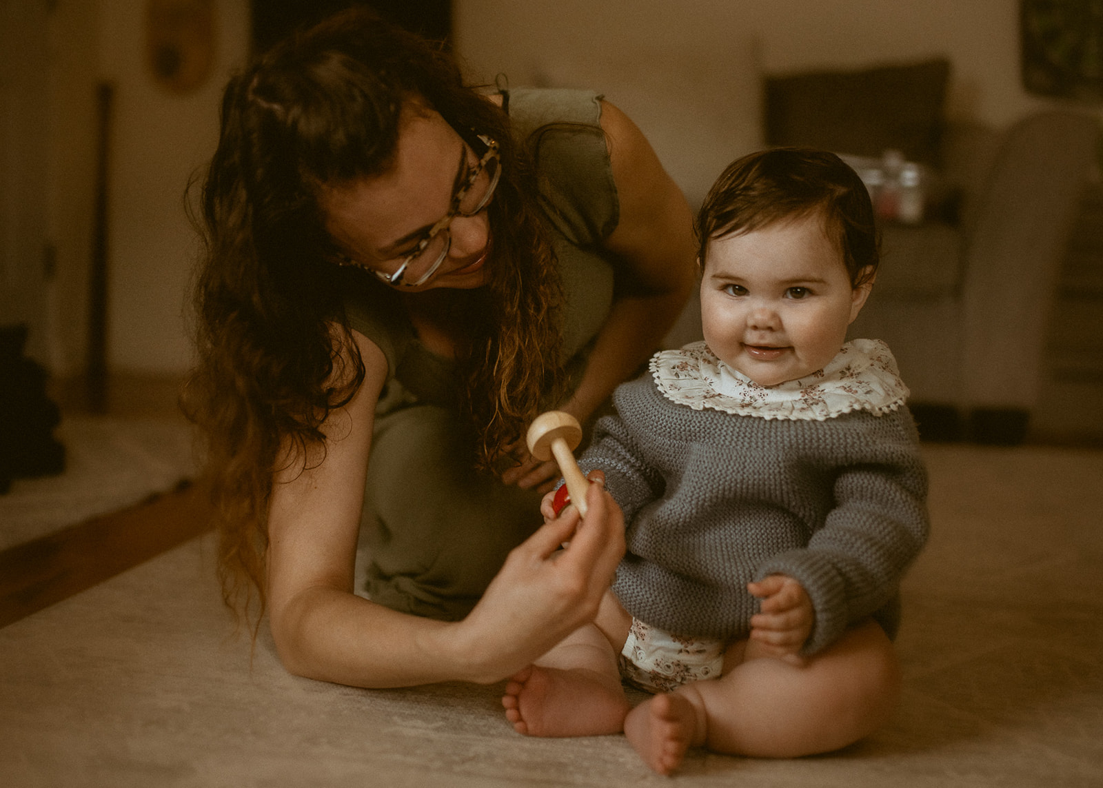A happy baby plays with mom with a wooden rattle in the living room in a blue knit sweater before visiting Roots Acro Sports