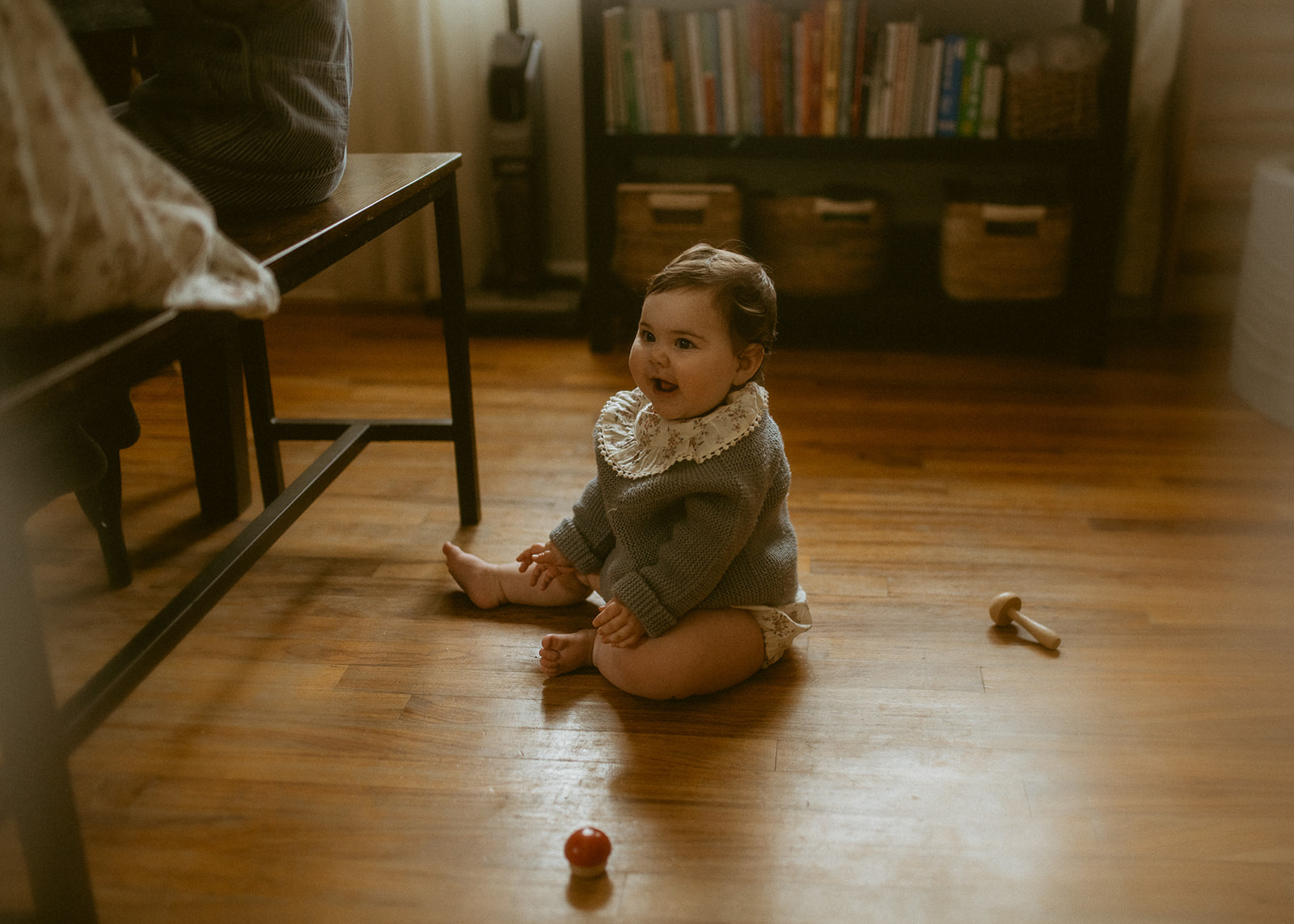 A happy baby girl in a knit sweater smiles while sitting in the dining room with wooden toys before tubing in Missoula