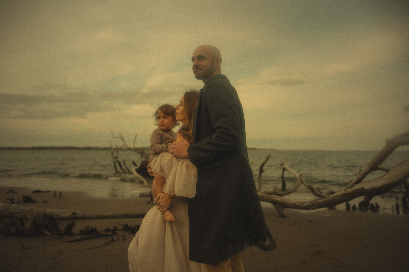 Happy mom and dad admire the water on a beach surrounded by driftwood holding their toddler