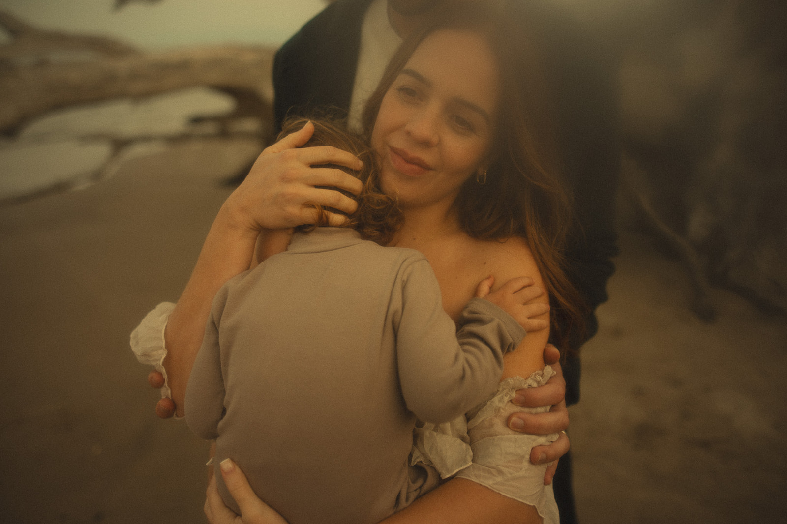 A smiling mother cradles her toddler against her shoulder while walking on a beach in a white dress after a trip with Zootown Surfers