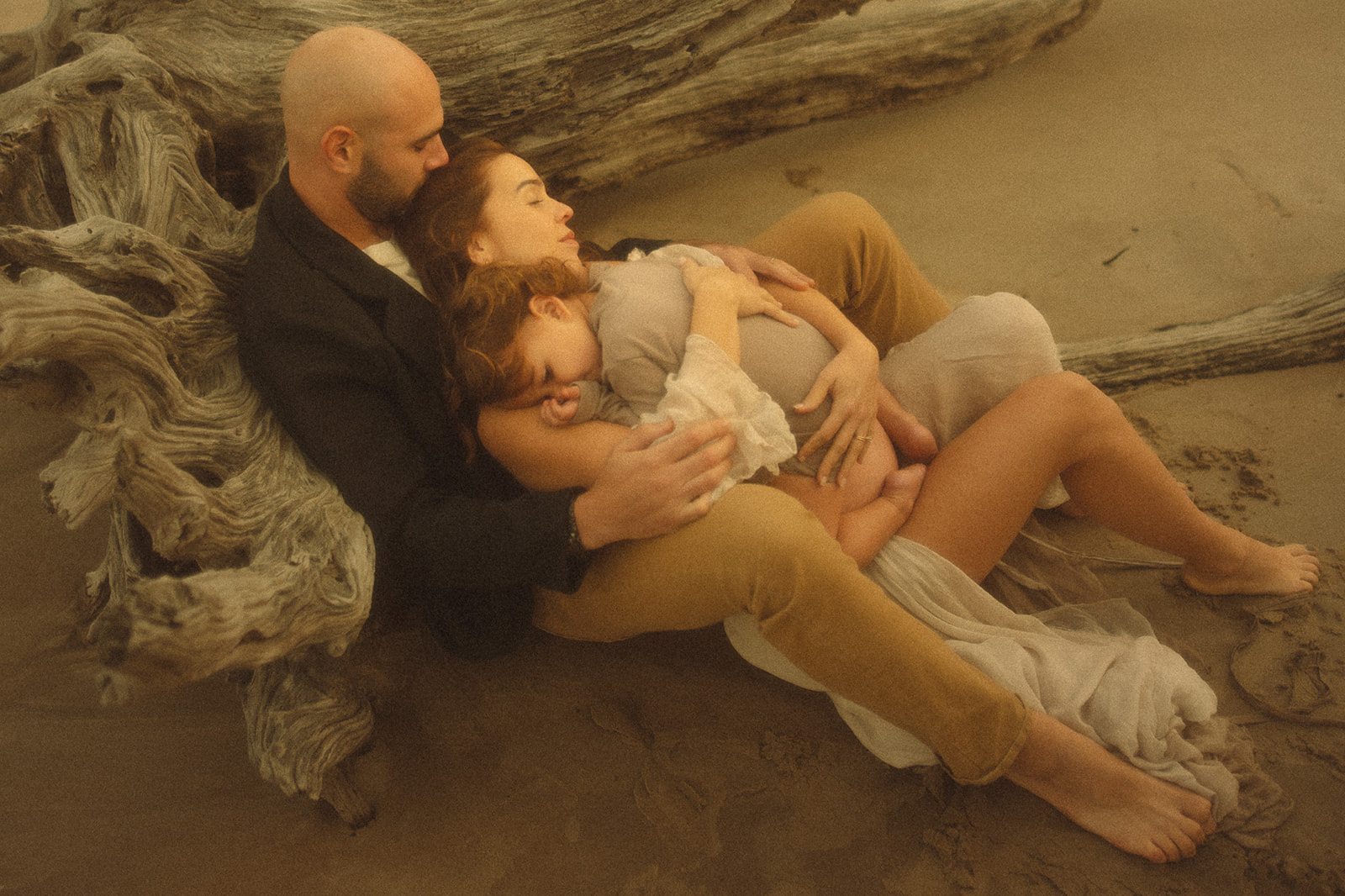 A mother and toddler snuggle in the lap of dad leaning on driftwood on a beach after a trip with Zootown Surfers