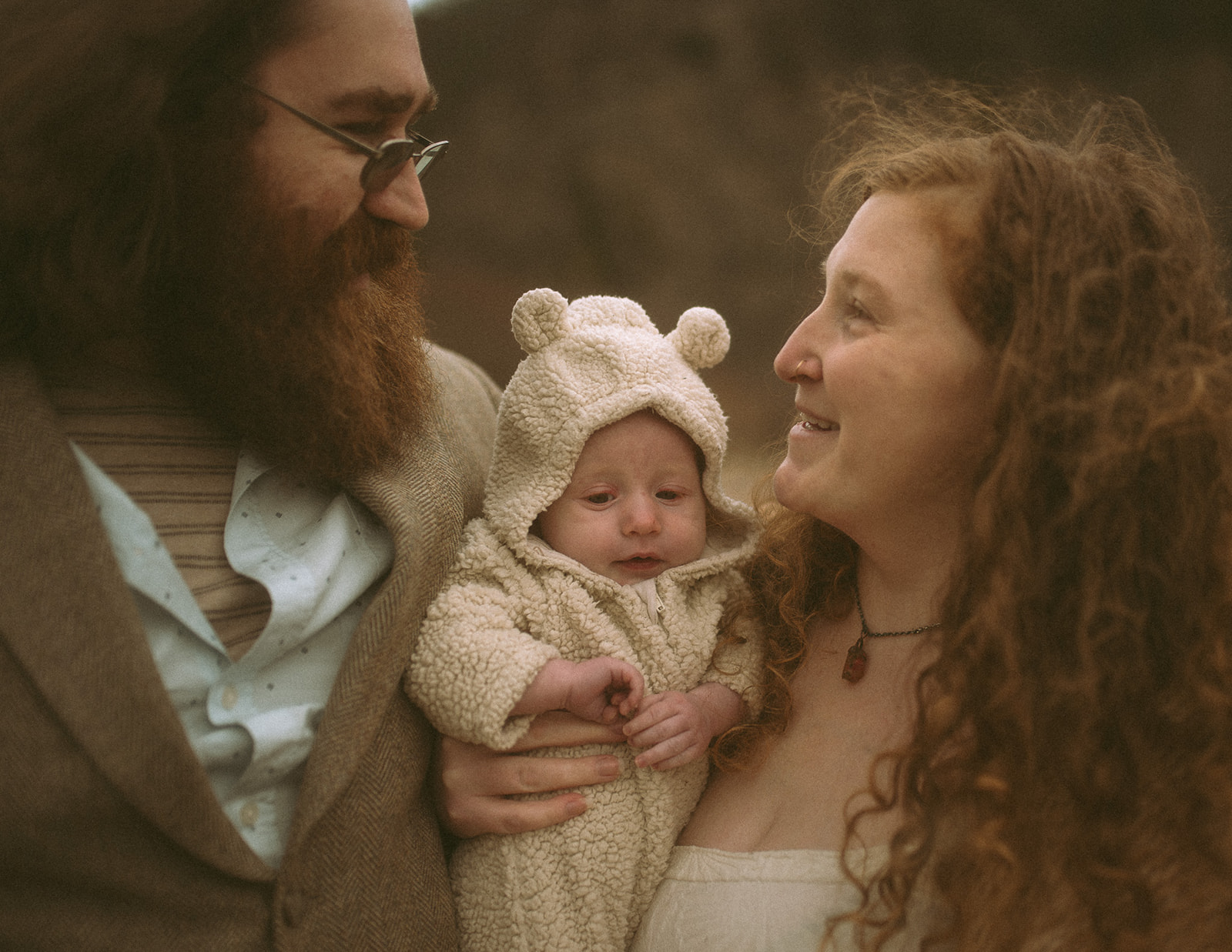 Happy new parents smile at each other while holding their newborn in a teddy bear onesie between them after visiting daycares in Hamilton, MT
