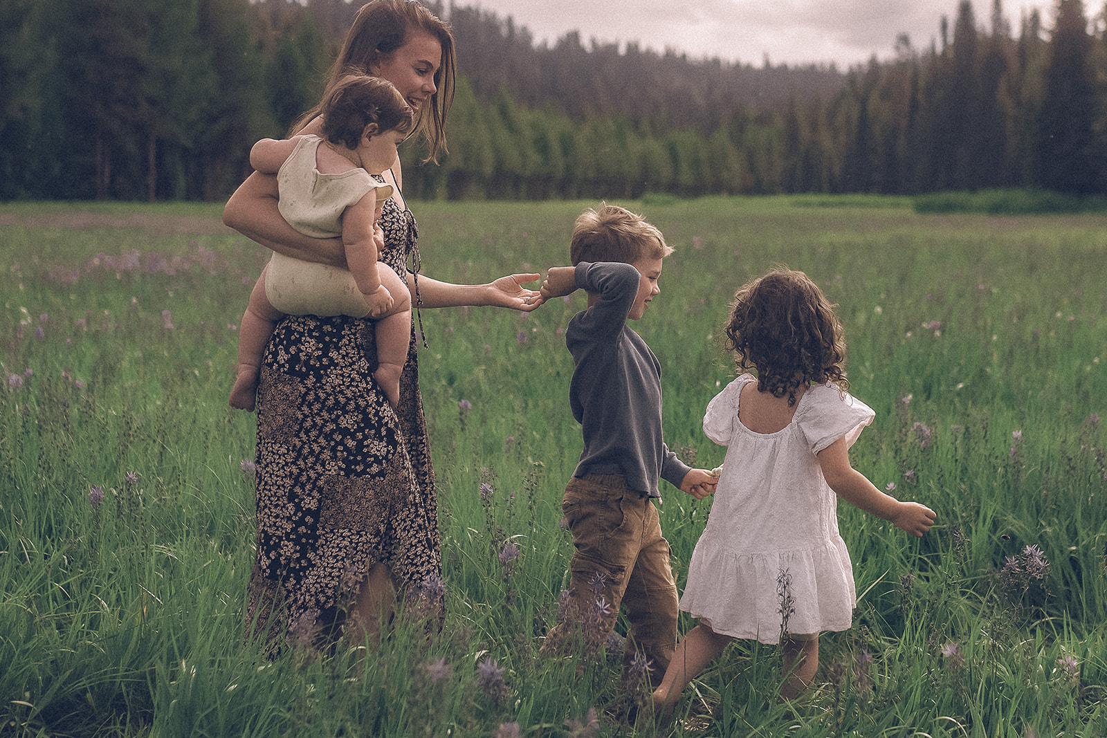 A happy mother walks with her two toddlers through a meadow of wildflowers with an infant on her hip in a floral print dress