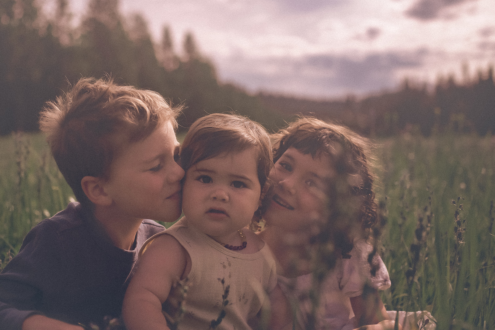 Three young siblings smile with faces together in a meadow of tall grasses after exploring Endvr Missoula