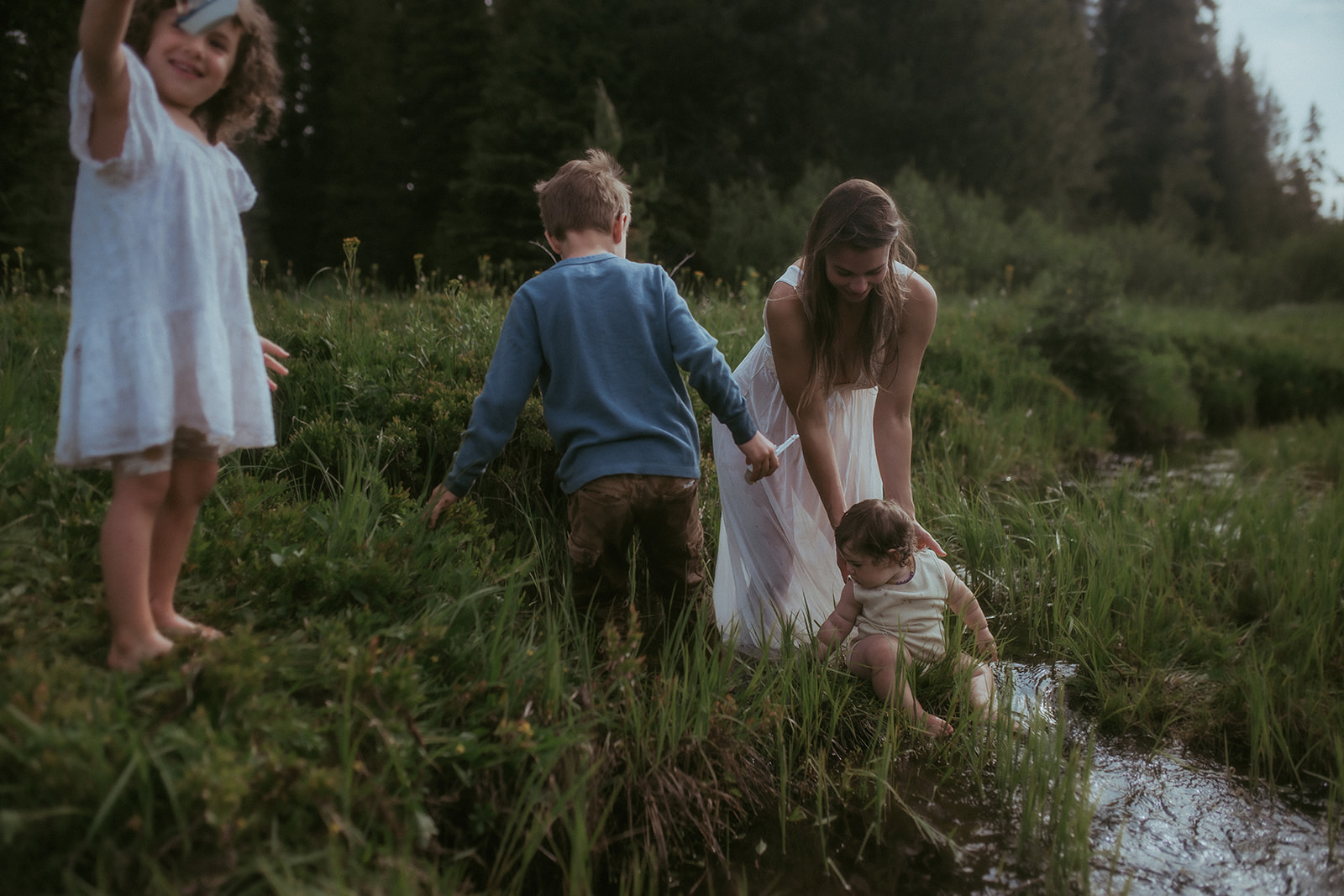 A mother helps her three young children explore a shallow creek in a white dress