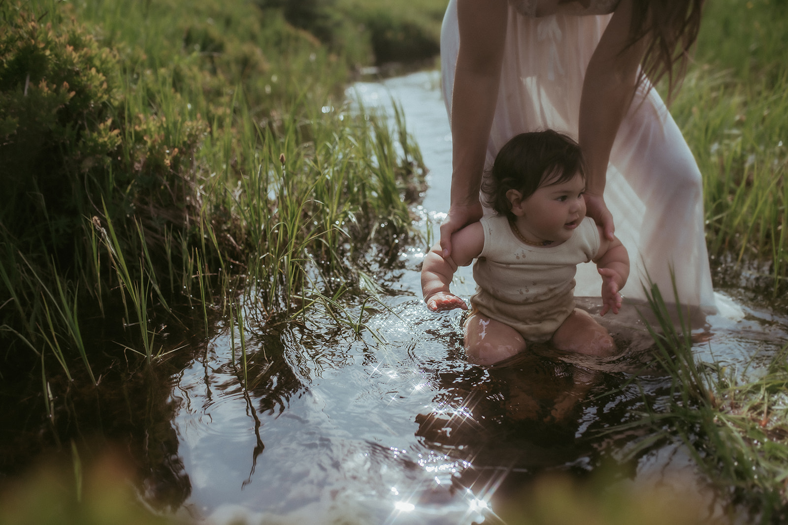 An infant girl in a white onesie is dipped into a shallow creek by mom in a white dress after visiting Ignite Creative School