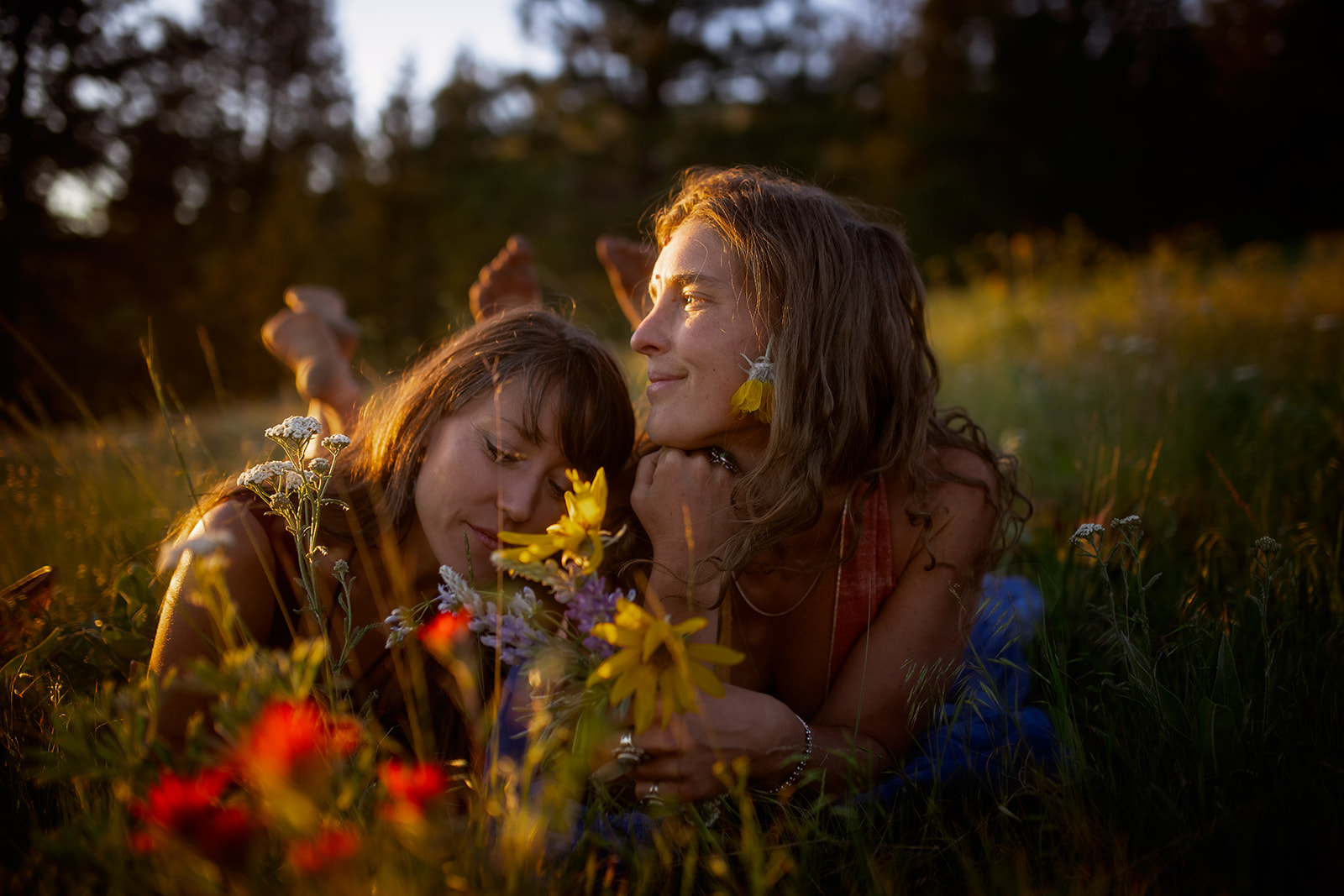 Two women snuggle in a grassy pasture surrounded by colorful wildflowers at sunset