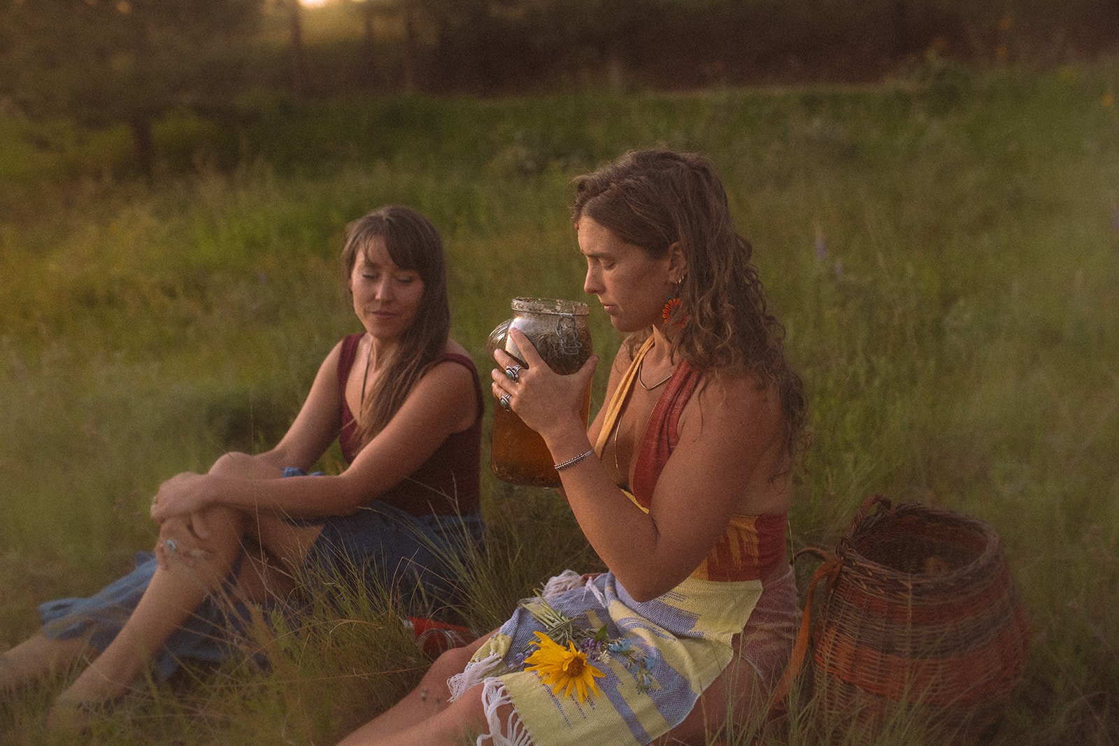 Two women sit on a grassy hillside at sunset in bright clothes as one smells a jar of tea with flowers in her lap after meeting with Somatic Connections