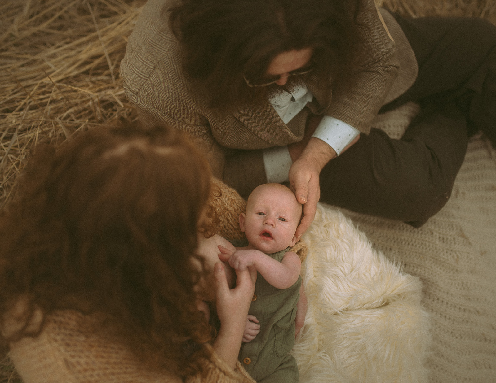 A newborn baby lays with eyes open in mom's lap as dad holds its head in a field of hay thanks to Tierra Madre Midwifery