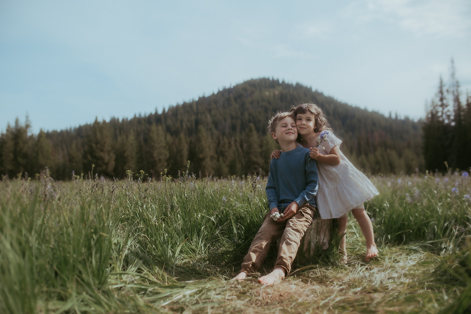 A young girl in a white dress hugs onto her bigger brother as he sits on a stump in a pasture