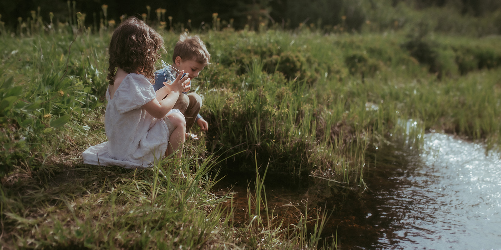 Toddler brother and sister sit on the bank of a small creek playing with toy boats after visiting Tinkergarten Missoula