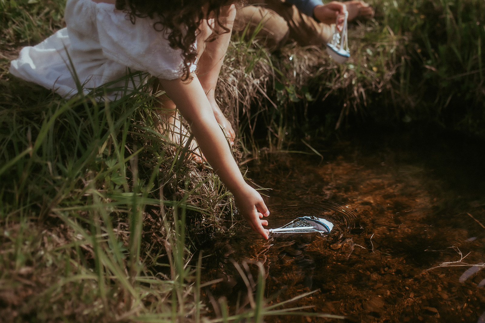 A young girl plays in a small creek with a toy boat in a white dress after visiting Tinkergarten Missoula
