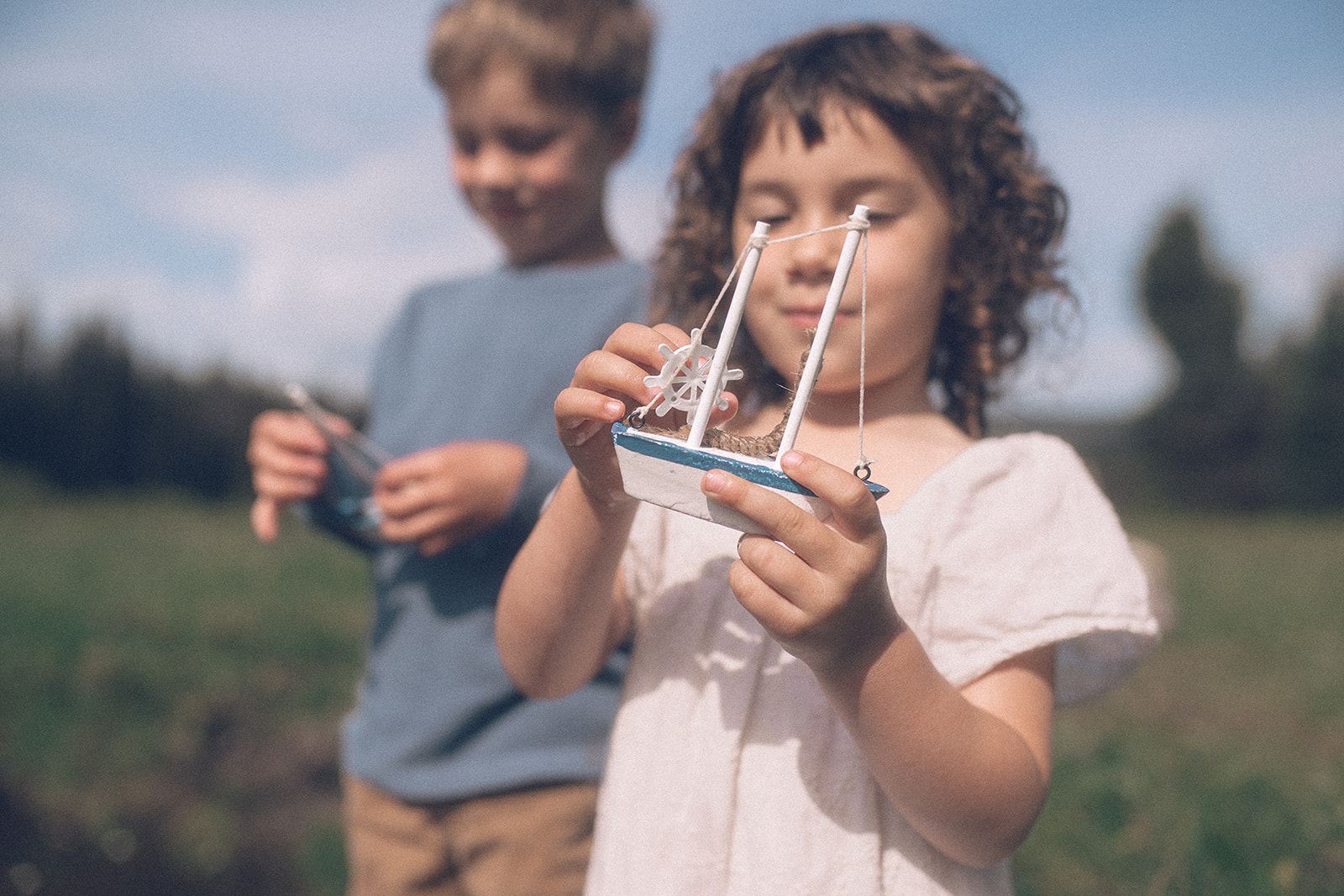 A happy young girl shows off her toy boat while wearing a white dress in a field with big brother
